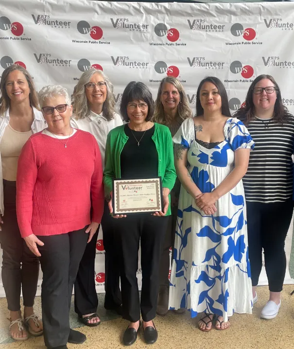 Group of 7 female volunteers and staff smiling and holding a framed award